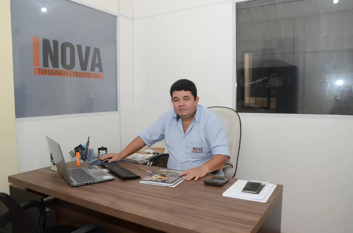 Profissional da Inova no escritório, sentado à mesa com notebook, com a marca Inova Topografia e Projeto na parede